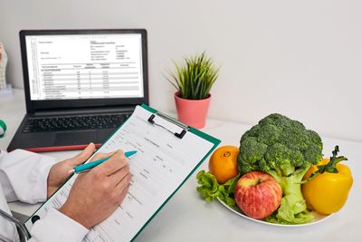 A person in a white coat fills out a patient form beside fresh fruits and vegetables.