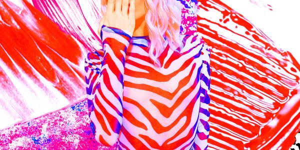 Person in vibrant zebra-patterned outfit and blue cap covers face against colorful abstract background.