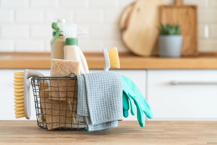 Basket of cleaning supplies including brushes, gloves, spray bottles, and cloths on a kitchen counter.