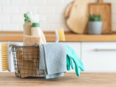 Cleaning supplies including brushes, gloves, and cloths in a wire basket on a wooden table.
