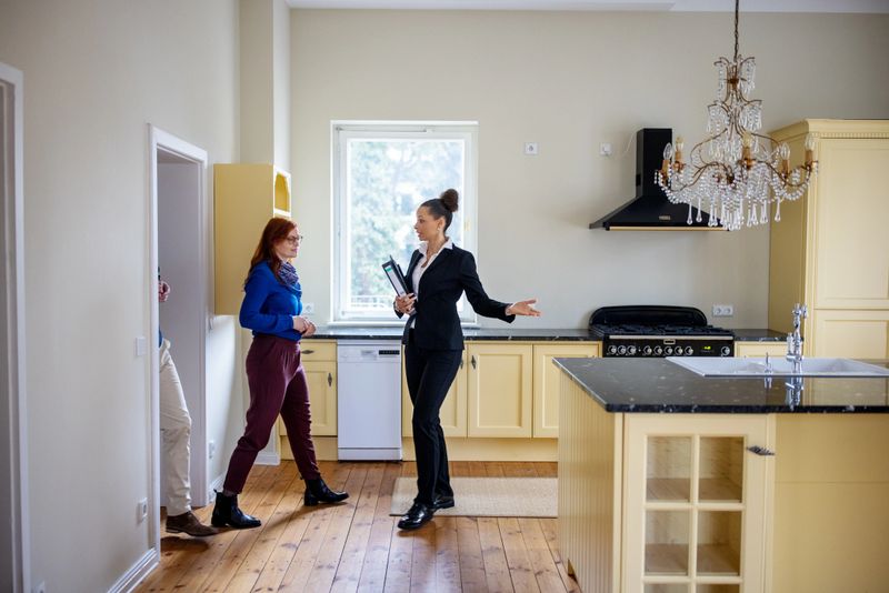 Female Real Estate Agent gesturing to customers at empty new apartment. Couple is entering kitchen. Saleswoman is showing home to man and woman.