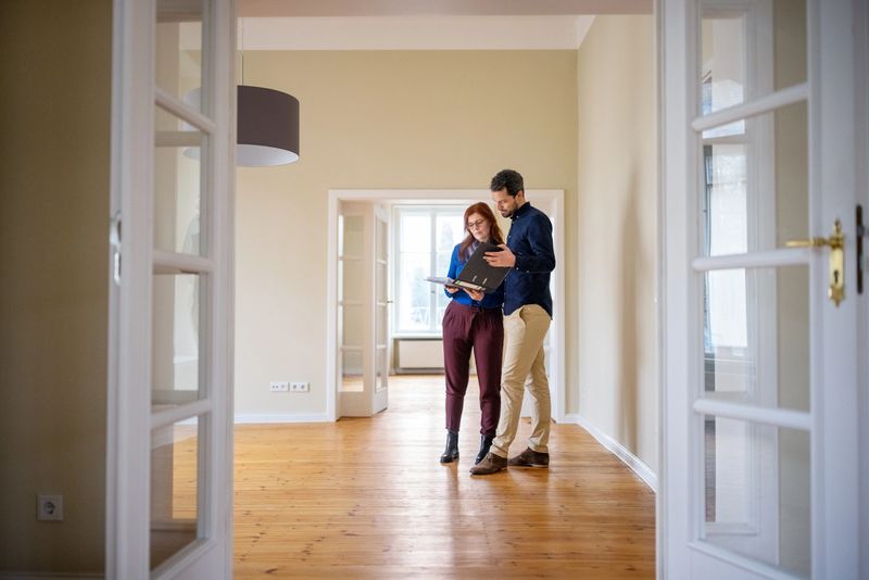 Full length of couple analyzing document while standing at empty new apartment. Man and woman are discussing together. They are seen through doorway.