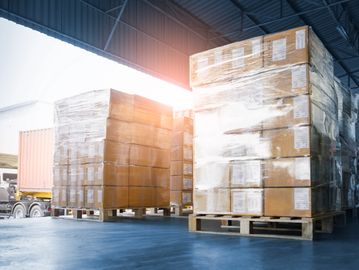 Stacked pallets of shrink-wrapped boxes in a warehouse with a truck nearby.