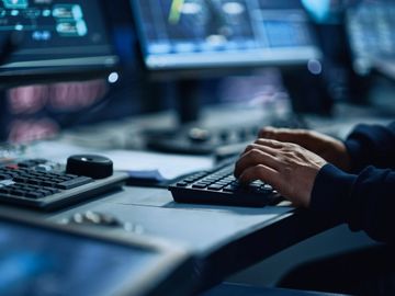 Person typing on a keyboard in a dimly lit, tech-heavy workspace.