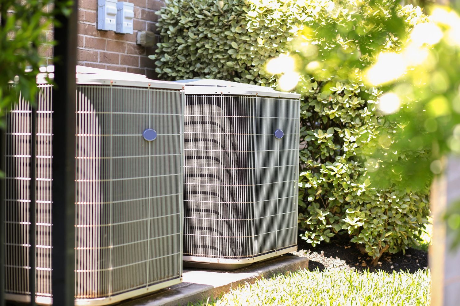 Two outdoor air conditioning units beside a brick wall with greenery.