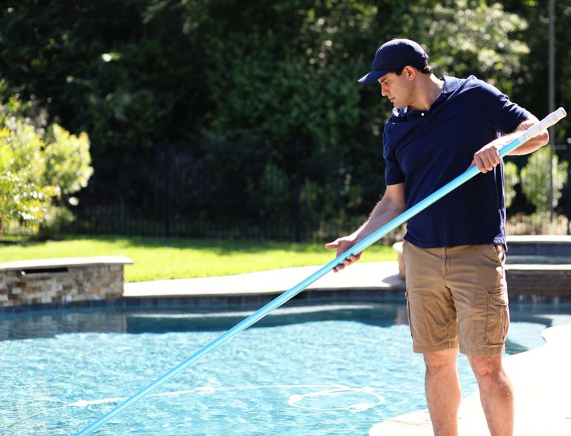 Latin descent man works for local swimming pool cleaning and repair service.  He uses net to clean out pool for summer at home of customer.