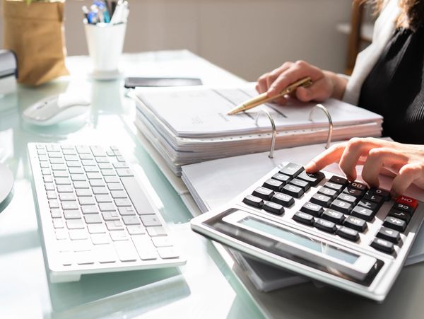 Person using a calculator and pen to review documents at a desk with a computer.