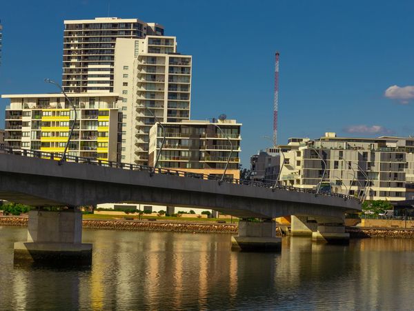 Concrete bridge over calm water with modern buildings in the background under a clear blue sky.