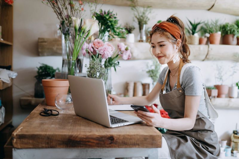 Asian female florist, owner of small business flower shop, using smartphone while working on laptop against flowers and plants. Checking stocks, taking customers orders, selling products online. Daily routine of running a small business with technology