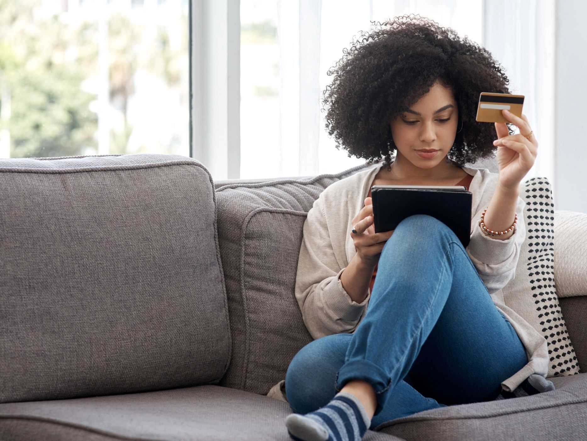 Young woman shopping online using a tablet and credit card on a couch.