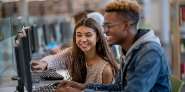Two students smiling and working together on a computer in a library.