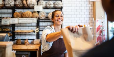 Smiling baker handing a paper bag to a customer in a bakery.