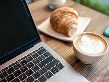 Laptop, cappuccino, and croissant on a wooden table.