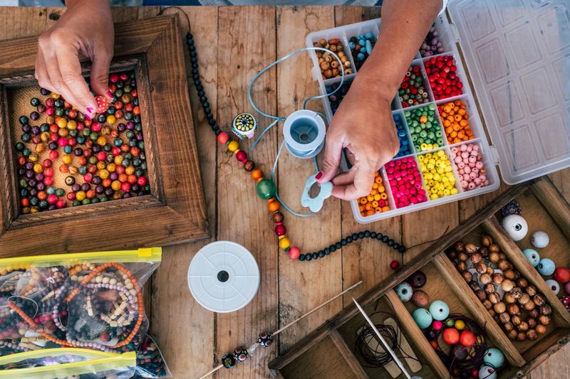 Making off handmade jewellery. Box with beads on old wooden table. Top view with woman hands - tutorial to learn how to make bracelets and jewellery online and alternative home job