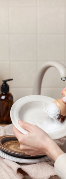Hands washing a white plate with a brush and soap foam near a kitchen sink.