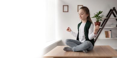 Young girl meditating cross-legged on a table near a window.