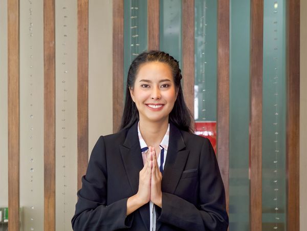 Smiling woman in business attire greeting with a traditional Thai wai gesture.