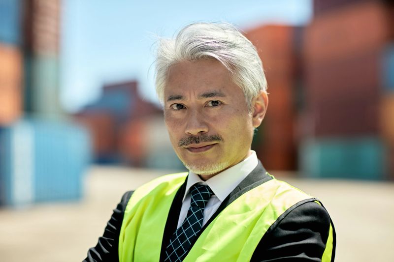 Three-quarter front view of man wearing reflective vest over suit and standing amidst cargo containers looking at camera with serious expression.