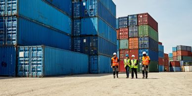 Workers in safety gear walk near stacked shipping containers.