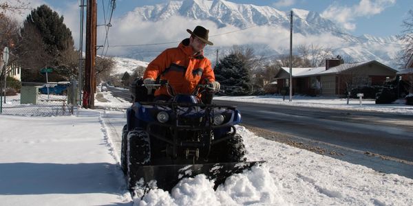 Man in orange jacket snow plowing a sidewalk with mountains in the background.