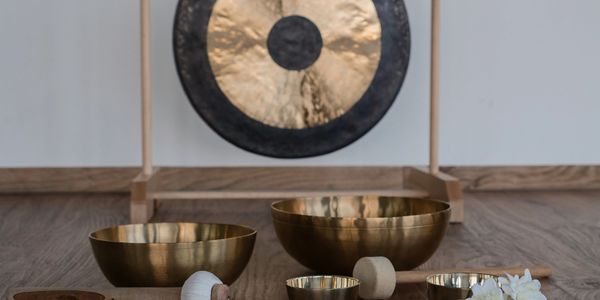 Meditation setup with golden singing bowls, mallets, a gong, and white flowers on wooden floor.