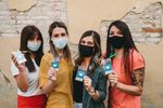 Four young women wearing masks holding up ice cream cones outdoors.