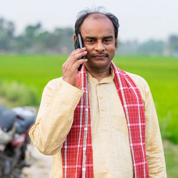 Man in traditional attire talking on phone in a rural setting.