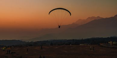 A paraglider soars over a sunset-lit landscape with mountains in the background.