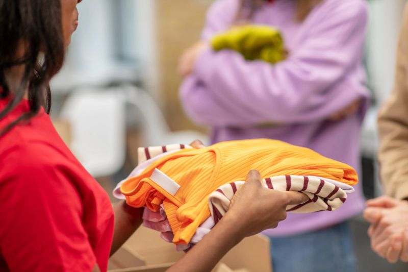 Help, clothes. Dark-skinned girl volunteer in red tshirt holding offering clothes for those in need of charity organization