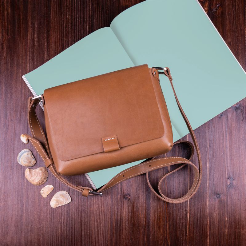 Brown women's cross body bag with a magazine and shells is lying on a brown wooden table