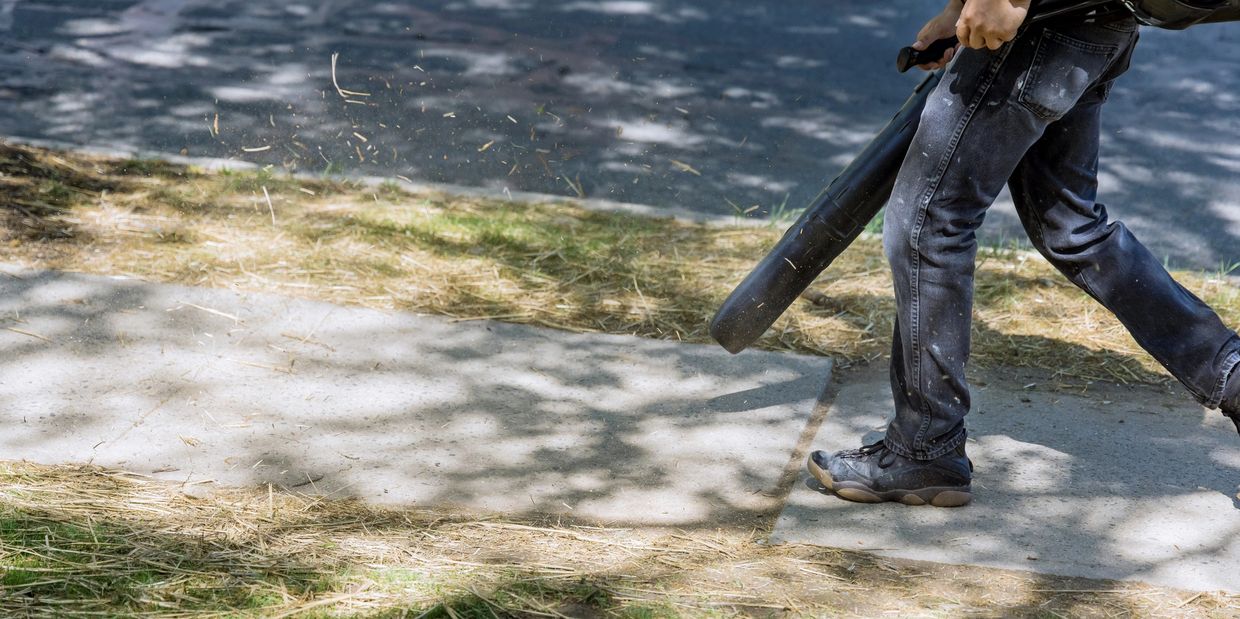 man using a leaf blower to clean off a sidewalk after mowing a lawn.