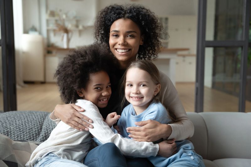 Family portrait of African American mom sit on sofa at home relax with multiethnic daughters. Smiling biracial young mother rest on couch with diverse multiracial girls children. Adoption concept.