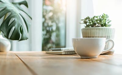 A white coffee cup on a wooden table with plants and a notebook in soft sunlight.