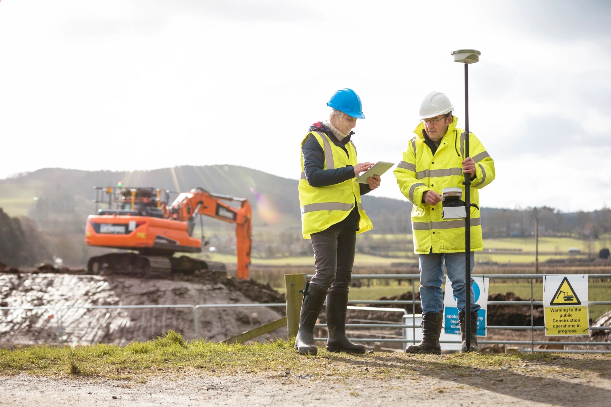 Two construction workers in safety gear using surveying equipment.