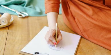 Person sketching on paper with pencil while sitting on wooden floor.