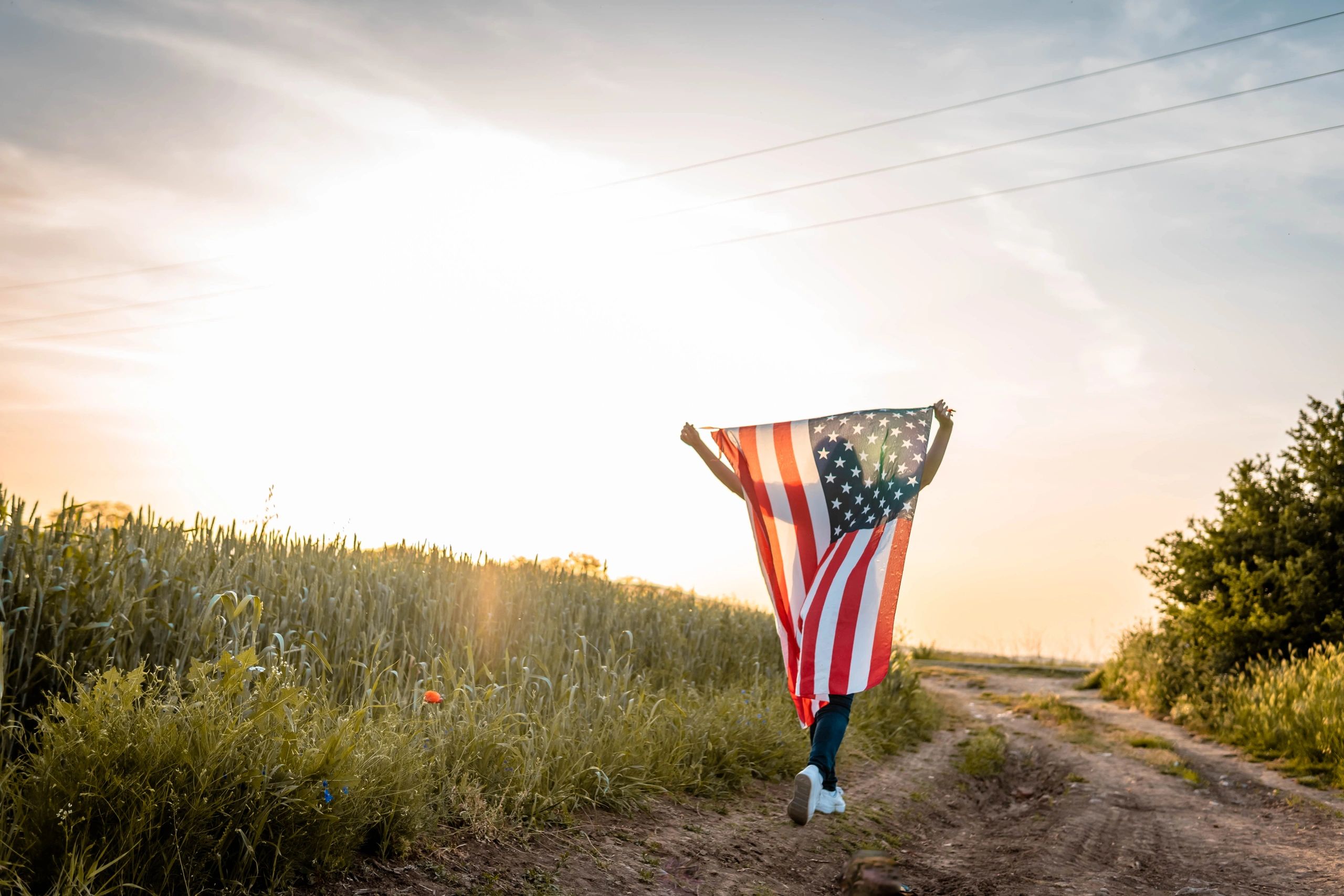 Person holding an American flag walking on a dirt path at sunset.