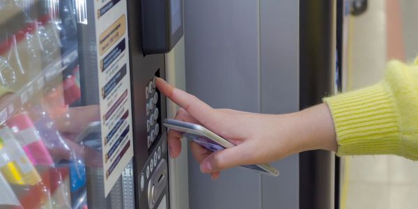 Hand pressing a button on a vending machine while holding a phone.