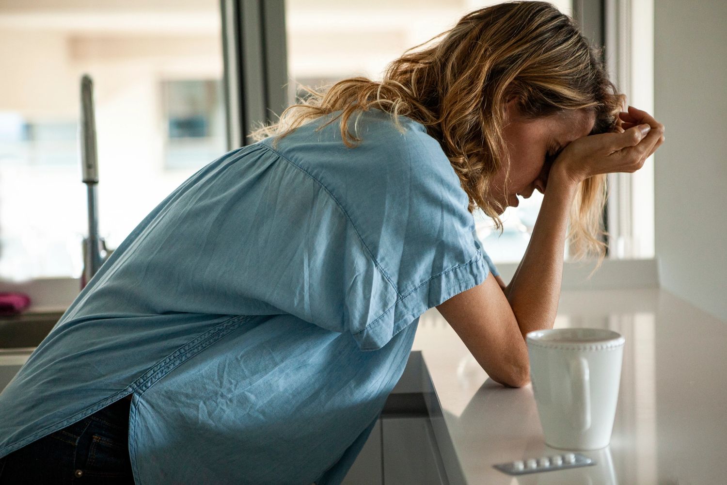 Frustrated woman with headache is holding her head and leaning on her kitchen counter. 