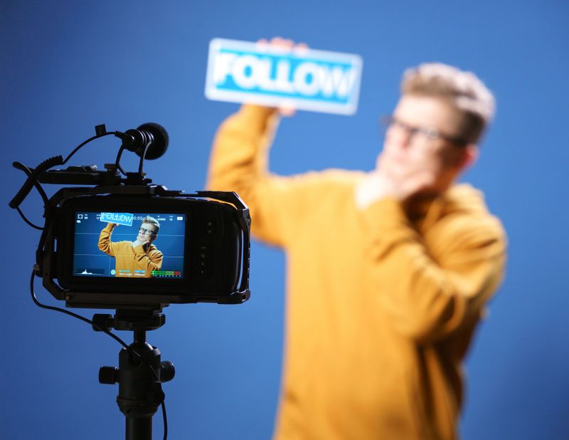 A photo from the filming of a vlogger who is broadcasting live on the Internet on his channel using a professional digital camera. The vlogger advertises his social networks and asks viewers to subscribe to his video channel, for this he shows a sign "follow"