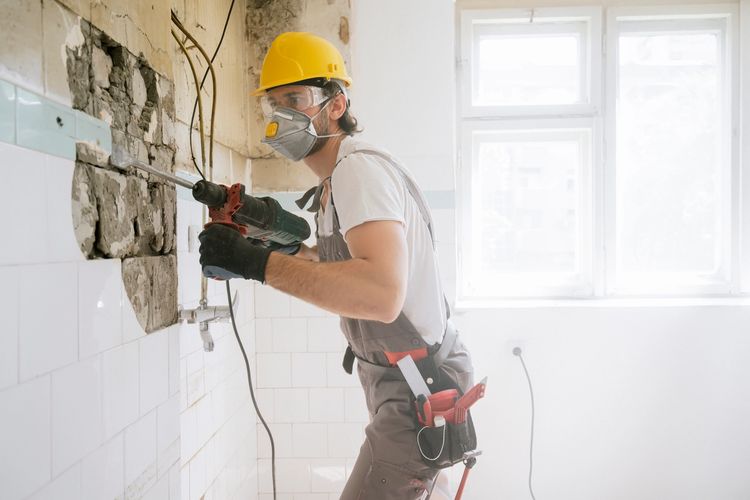 Construction worker drilling into tiled wall wearing safety gear.
