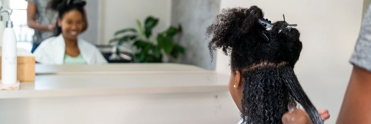A woman gets her curly hair styled at a salon with a stylist working on the back.