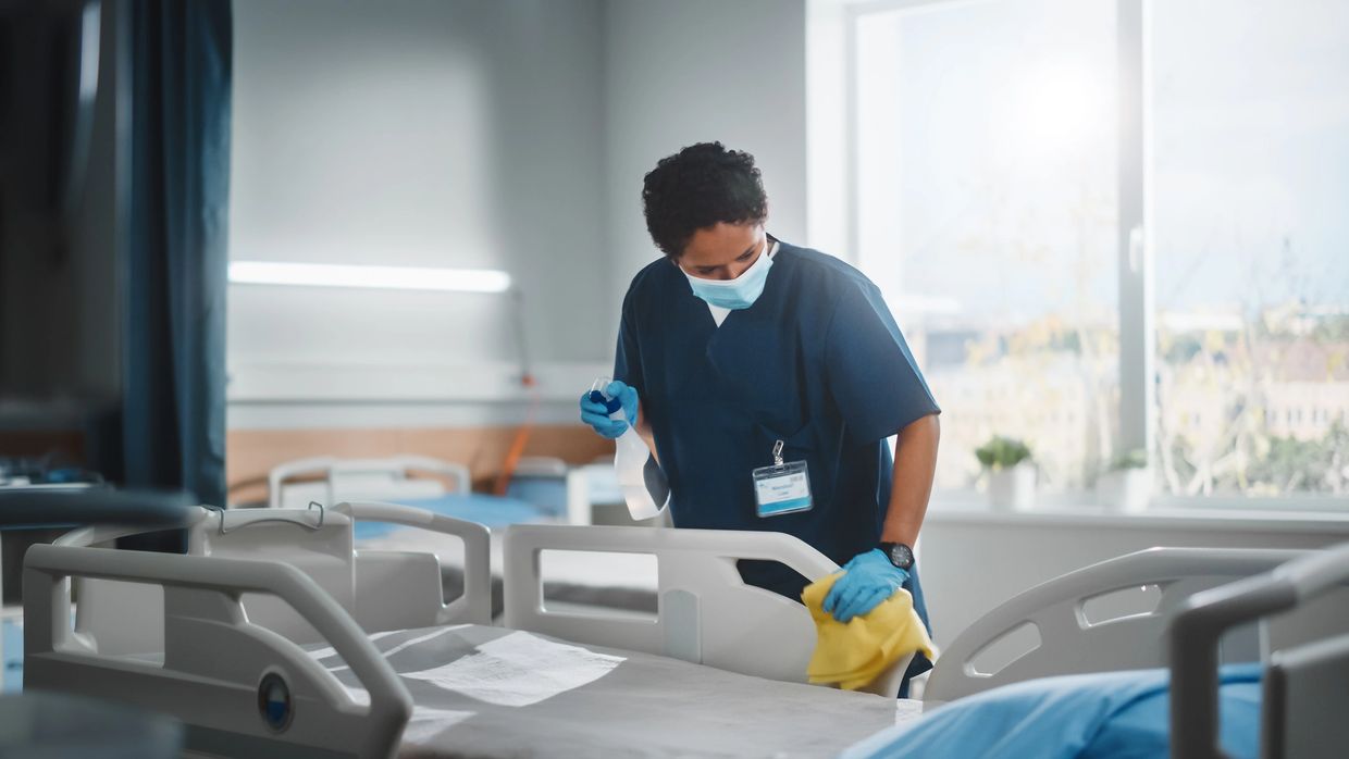 Healthcare worker disinfecting a hospital bed with cleaning spray and cloth.