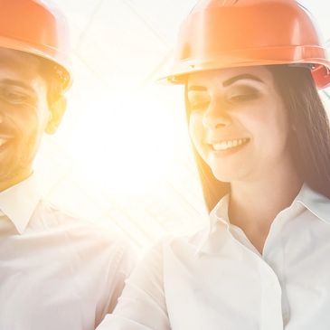 Two architects in orange helmets smiling and discussing plans indoors.