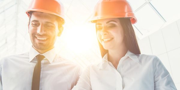 Two architects in orange helmets smiling and discussing plans indoors.