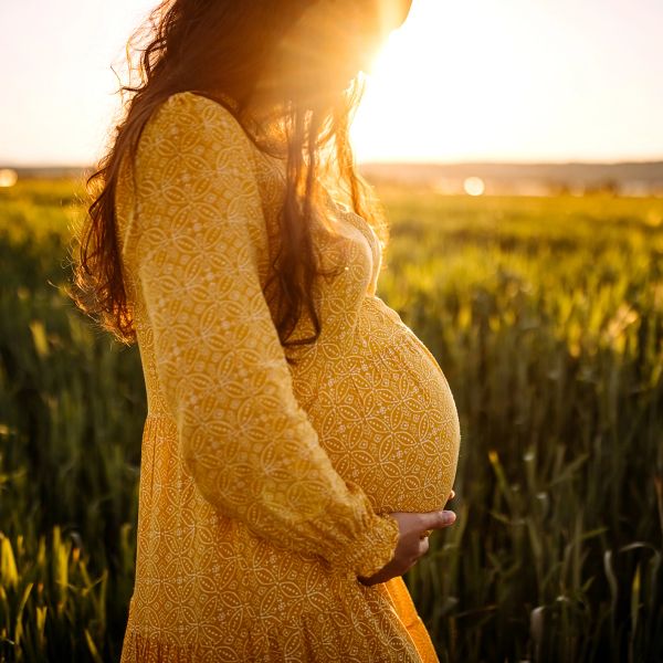 Pregnant woman in yellow dress standing in field at sunset.