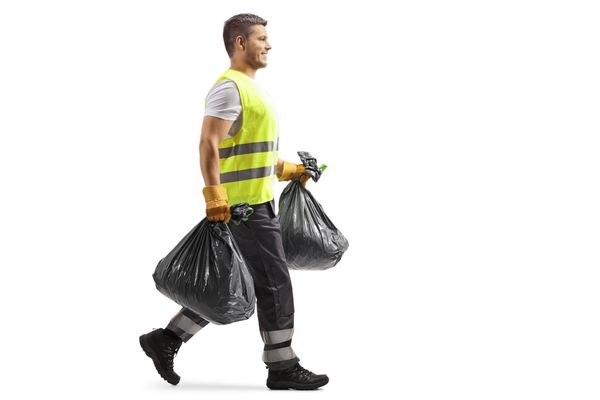 A sanitation worker carrying two black garbage bags, wearing a reflective vest and gloves.