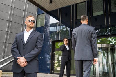 Three men in suits, two as bodyguards, one on phone, outside a modern building.