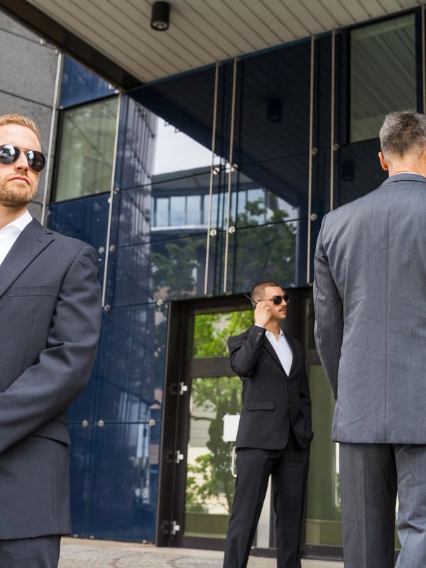 Three men in suits and sunglasses standing outside a modern building, appearing as security personnel.