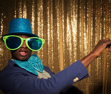 Man wearing oversized green glasses, blue hat, and bow tie posing playfully.