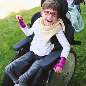 Happy child in wheelchair with pink arm braces outside.